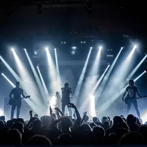 Ein Bild mit einer Bühne, Scheinwerfern, Künstlern auf der Bühne und Publikum davor.