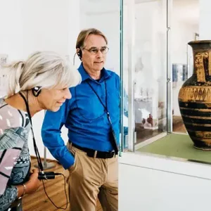 Bild von einem älteren Ehepaar im Museum, das sich mit Kopfhörern im Ohr interessiert ein Ausstellungsstück ansieht.