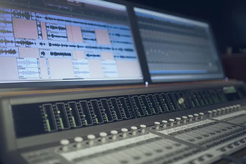 Ein Bild mit einem Mischpult mit einem Bildschirm darüber, auf welchem Audiospuren zu sehen sind.